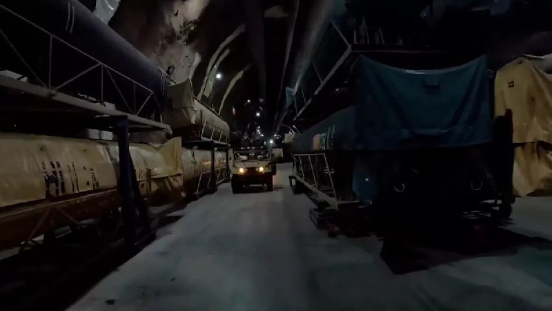 A small vehicle with headlights on drives down a dimly lit underground tunnel flanked by large heavy equipment and mining machinery.