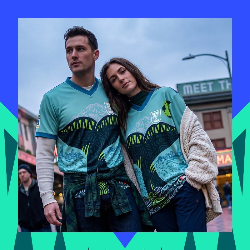 A man and woman wearing matching teal jerseys with shark-mouth and cityscape graphics stand close together outdoors, with a partially visible "MEET TH" sign in the background.