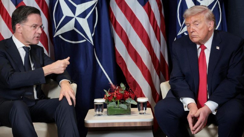 Two men in dark suits sit facing each other in armchairs, with the man on the left gesturing while speaking, against a backdrop of U.S. and NATO flags.