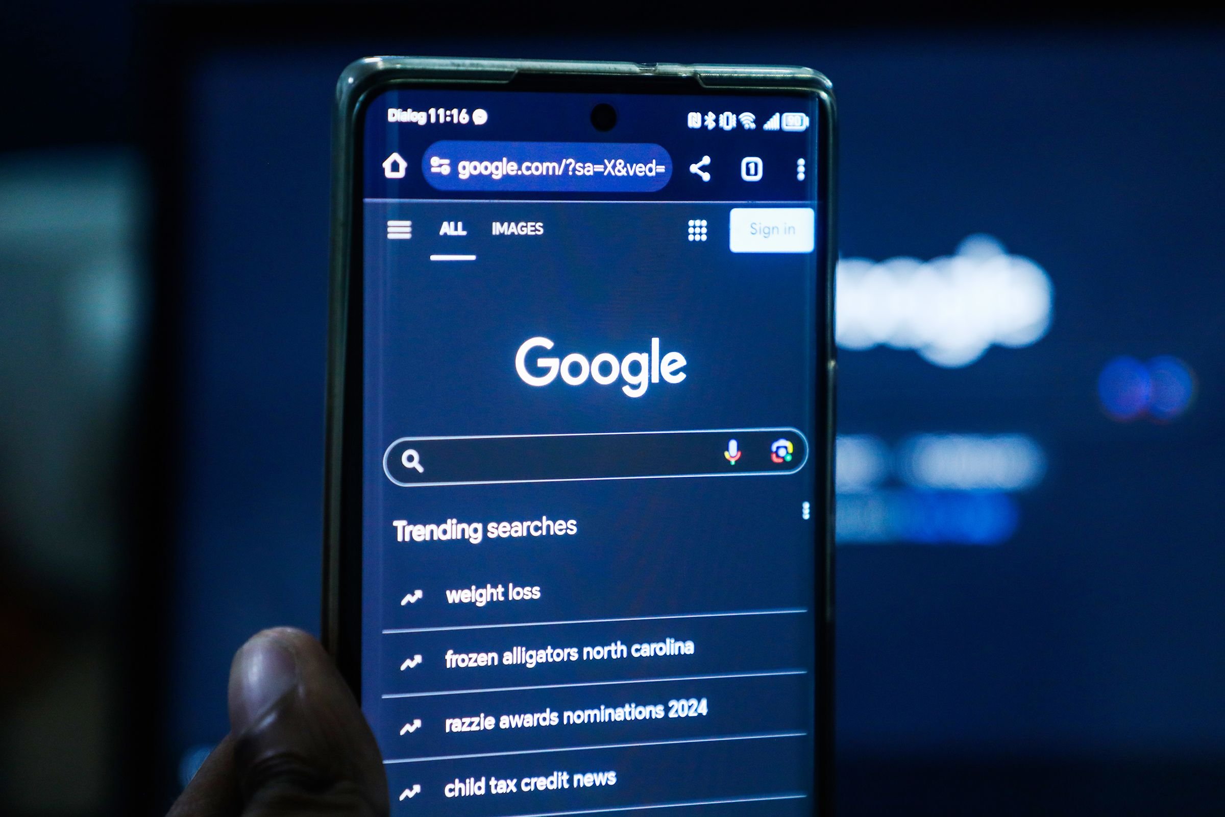 A hand holds a smartphone displaying the Google search homepage, with "Trending searches" including "weight loss" and "frozen alligators north carolina" visible on the screen.