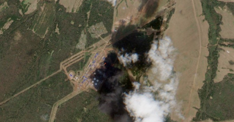 Aerial satellite image shows black smoke and visible flames rising from an industrial site in a rural, forested area, partially obscured by clouds.