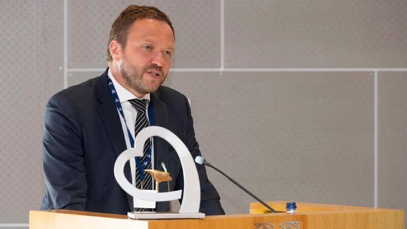 A bearded man in a dark suit and striped tie speaks at a wooden podium with a microphone, a glass of water, and a decorative silver award visible.