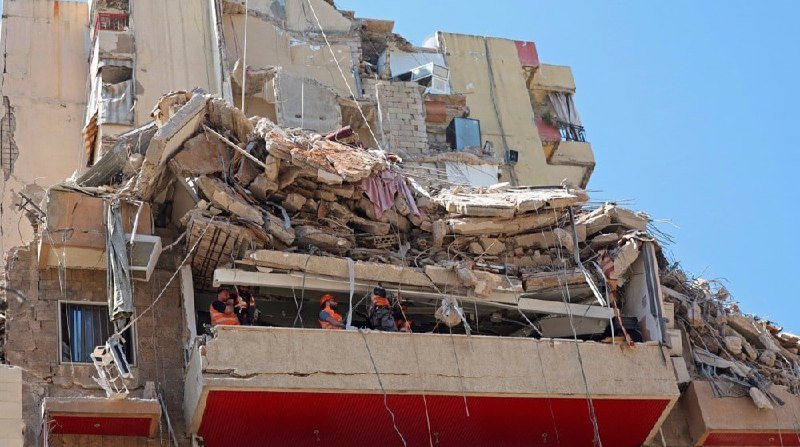 Rescue workers in orange vests search through the rubble of a partially collapsed multi-story building.