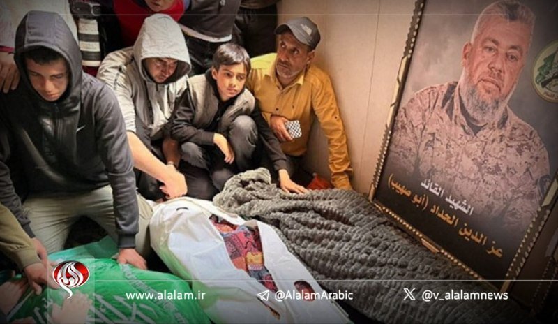 Mourners in hooded jackets gather around a body covered in a patterned cloth, beside a framed portrait bearing Arabic text, with an "Al-Alam" news watermark visible.