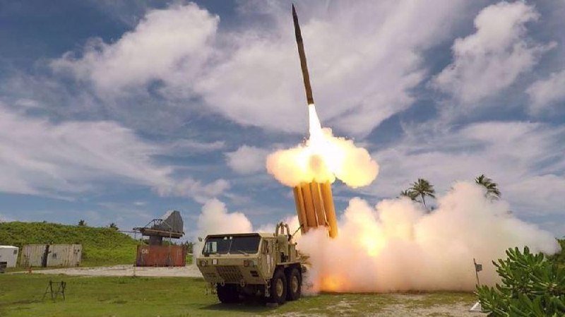A missile launches from a mobile military launcher vehicle, billowing flames and smoke, with a radar installation and shipping container visible nearby under a partly cloudy sky.