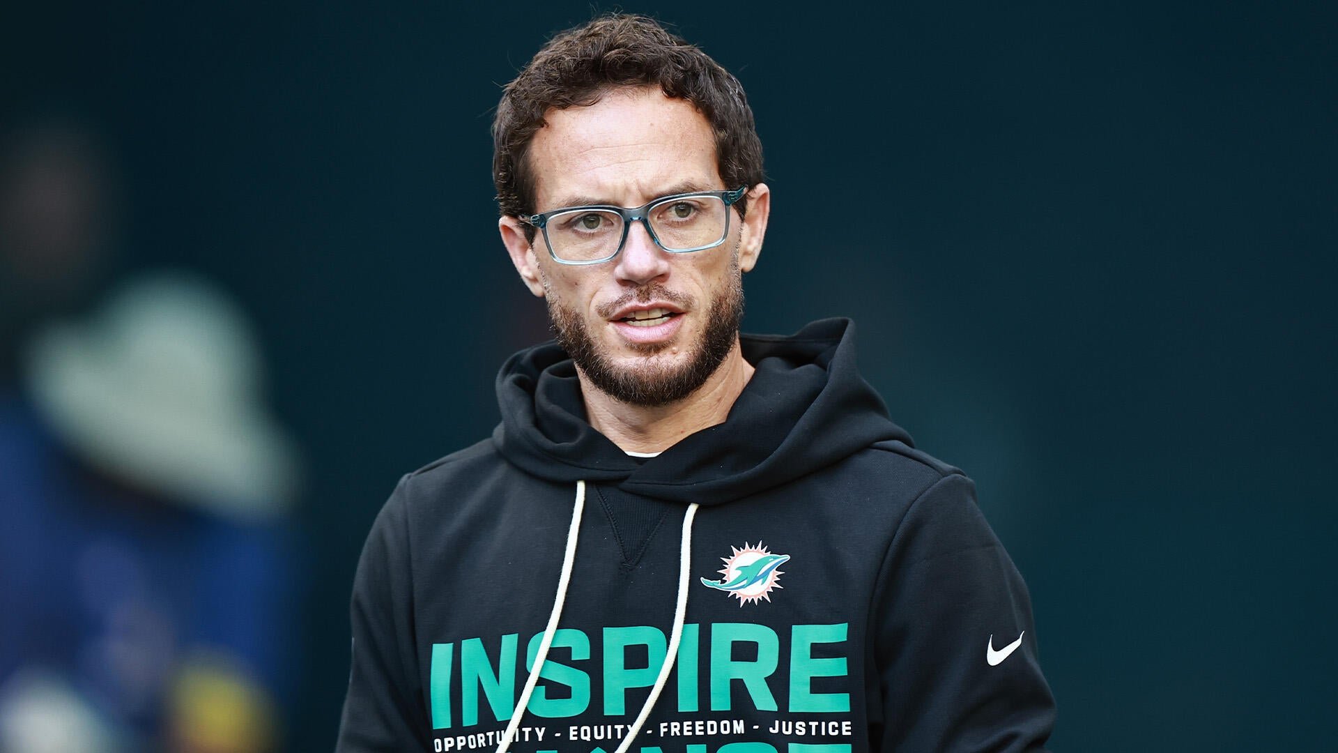A bearded man with glasses wears a black Miami Dolphins hoodie reading "INSPIRE" against a dark background.