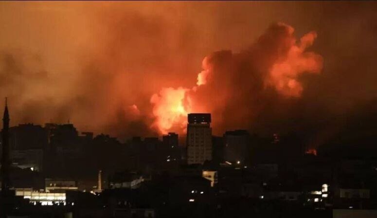 Large orange flames and thick smoke rise above a darkened city skyline at night, illuminating the clouds behind silhouetted buildings.