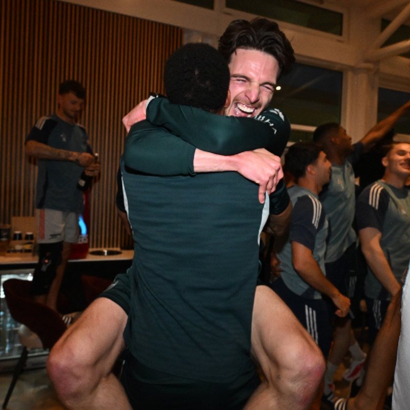 Two men in athletic gear embrace joyfully in a locker room, with one lifting the other, while teammates in matching uniforms celebrate in the background.