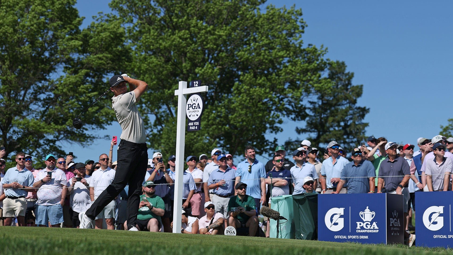 A golfer in a beige shirt and black pants swings a driver on a tee box while a large crowd of spectators watches near a "PGA of America" sign marking the 12th hole.