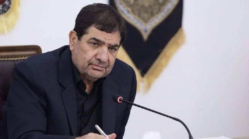 A man in a dark suit sits before a microphone, holding a pen, with decorative fabric wall hangings visible behind him.