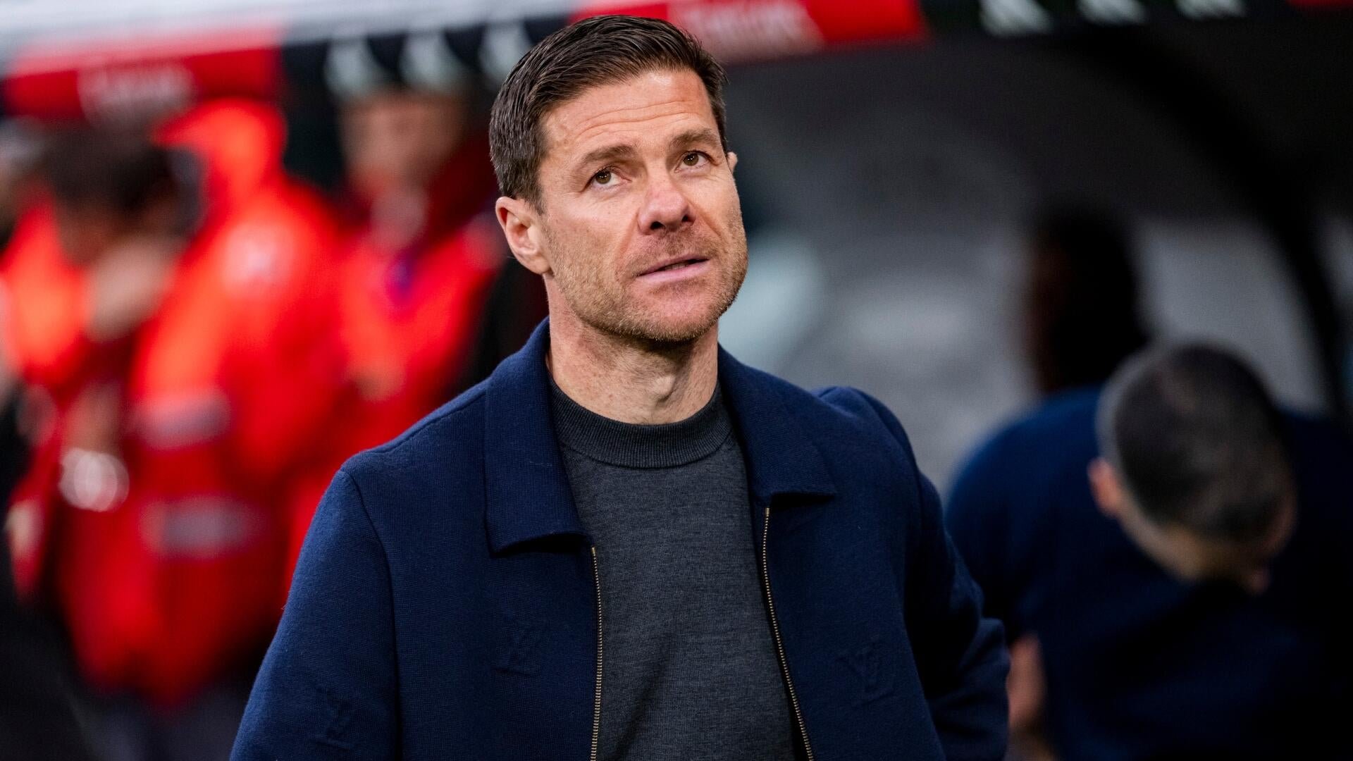 A man in a navy blue jacket and dark sweater looks upward, with blurred figures in red visible in the background.