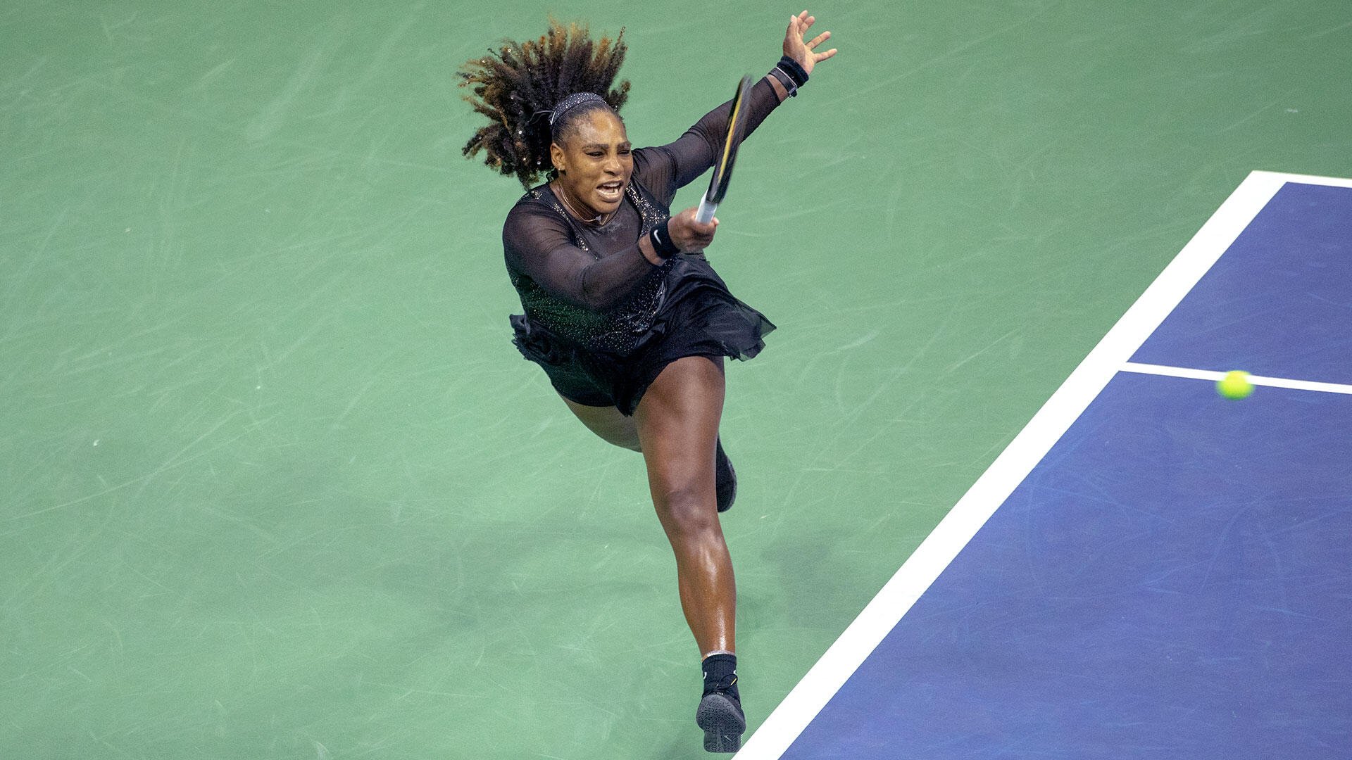 A female tennis player in a black outfit leaps to serve on a blue and green hard court, racket extended toward an incoming ball.