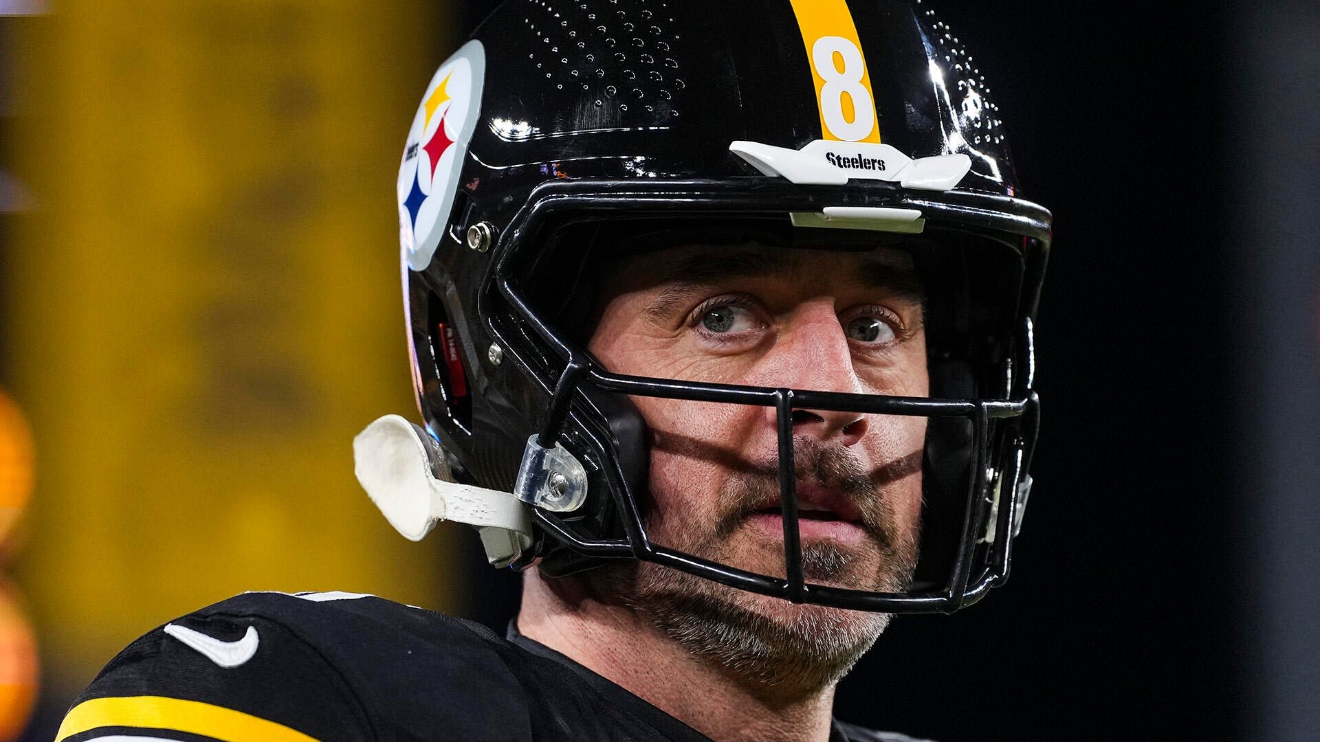 A football player wearing a black Pittsburgh Steelers helmet with the number 8 looks upward, standing against a blurred yellow and dark background.