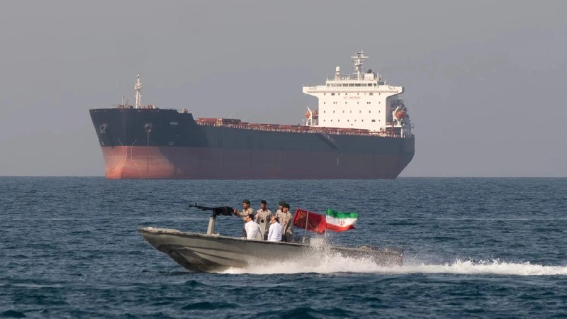 A small armed speedboat carrying several men and flying a green, white, and red flag speeds across the sea in front of a large red-and-black oil tanker.