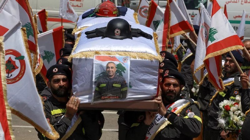 Uniformed personnel carry a flag-draped casket adorned with a helmet and a portrait photograph during a funeral procession surrounded by Lebanese flags.