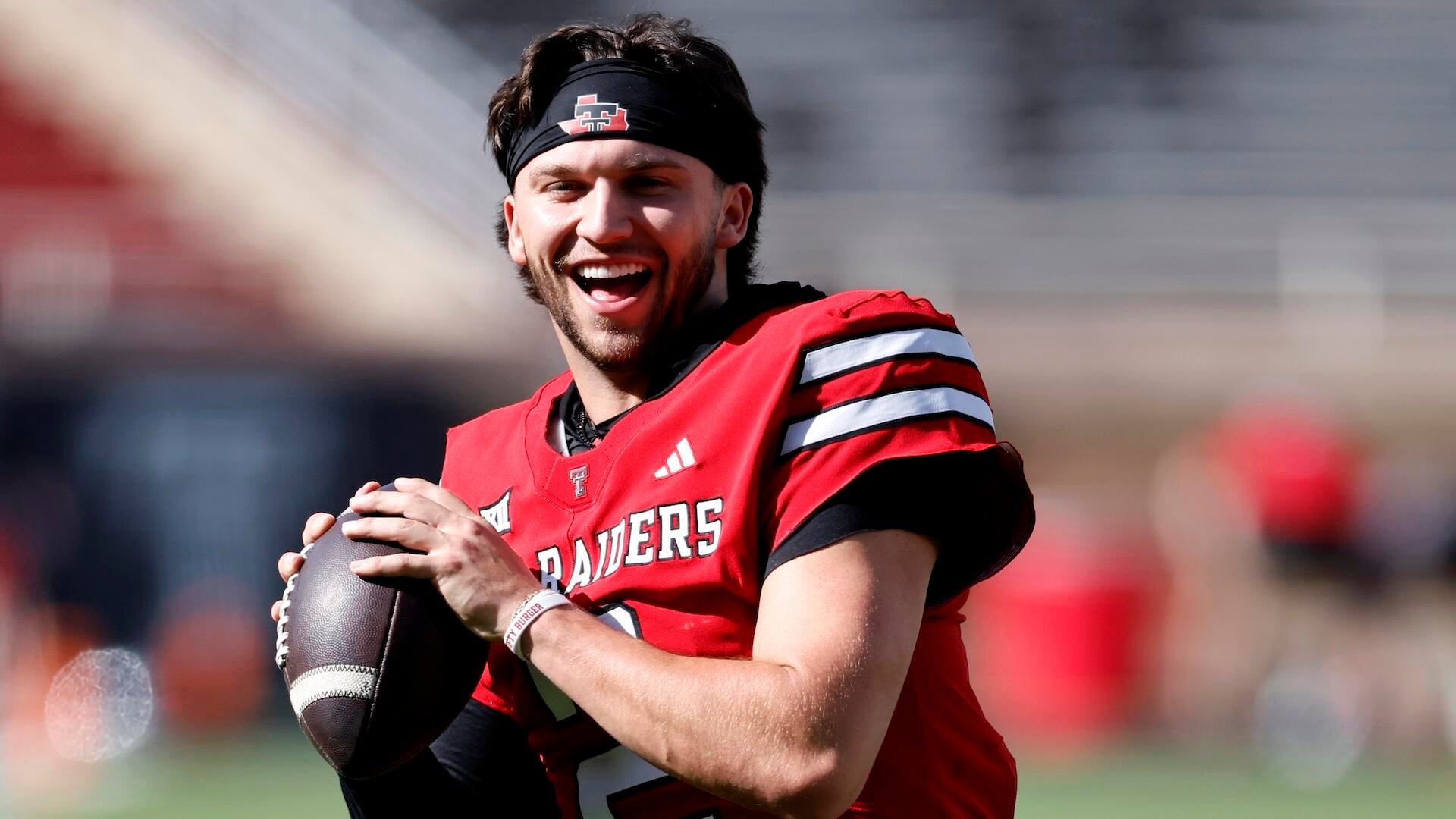 A smiling football player wearing a red "Raiders" jersey and a black "TT" headband holds a football on a sunlit field.