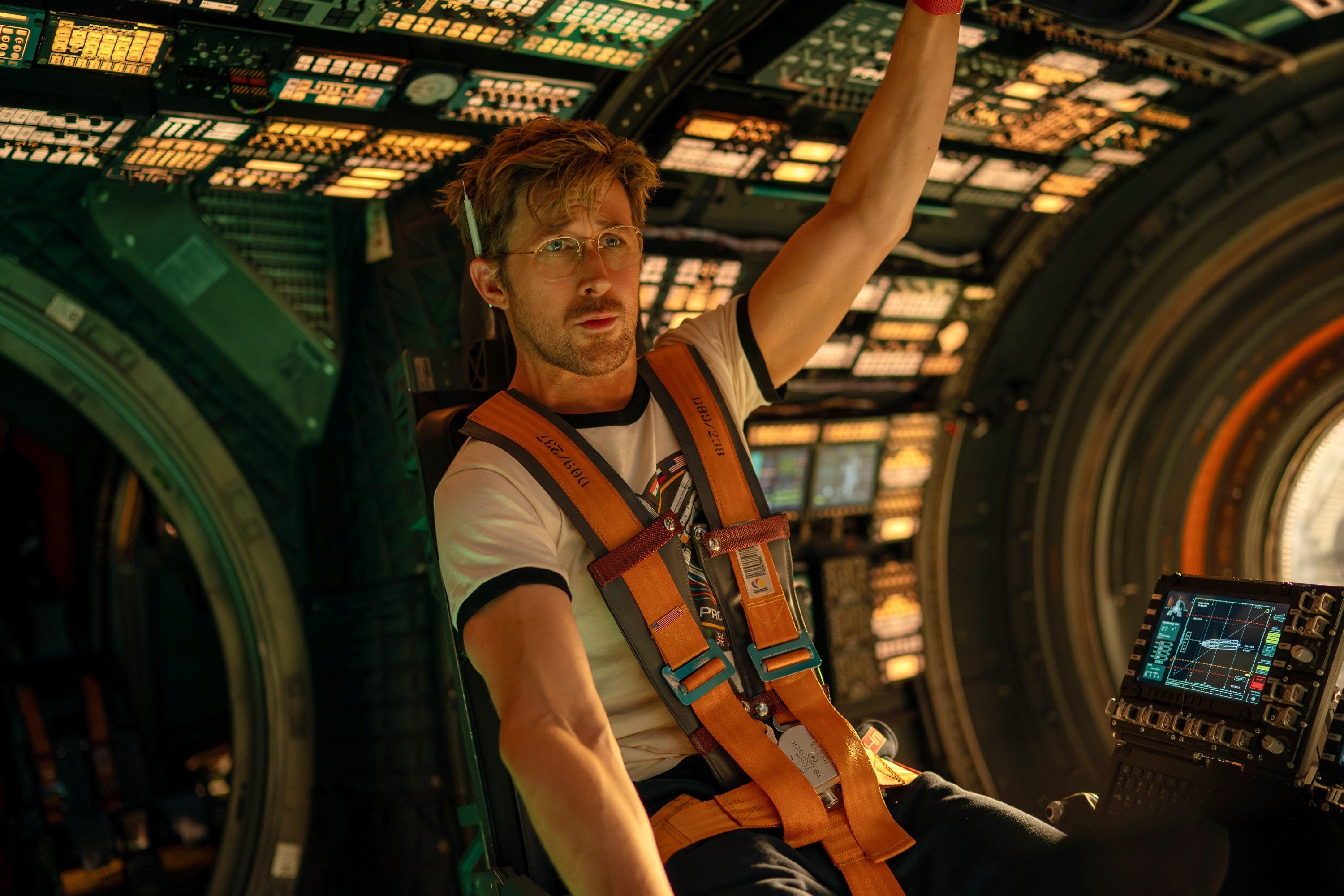 A bespectacled man in a graphic t-shirt and orange safety harness sits in a dimly lit spacecraft cockpit, reaching upward amid glowing control panels and monitors.