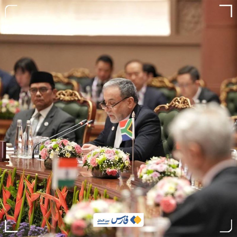 Officials in suits sit at a conference table adorned with floral arrangements and small South African and Indian flags, with microphones positioned before them.