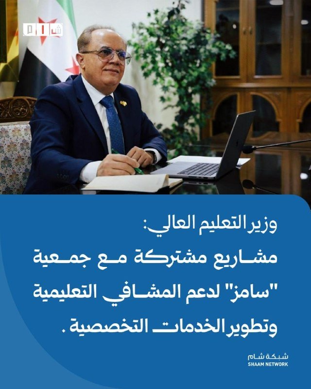 A bespectacled man in a suit writes at a desk with a laptop, set against a flag backdrop and wooden cabinets, with Arabic text overlay on a blue graphic banner.