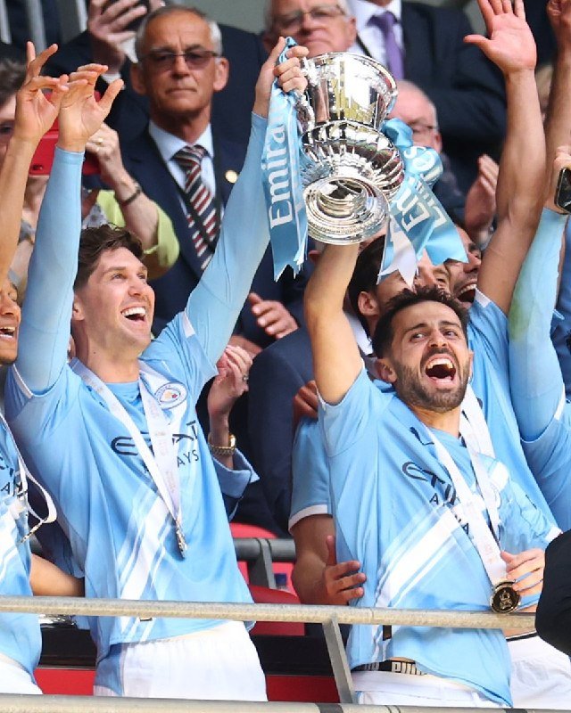 Two men in light blue jerseys and medals hoist a silver trophy with "Emirates" ribbons above their heads, celebrating amid a crowd of spectators.