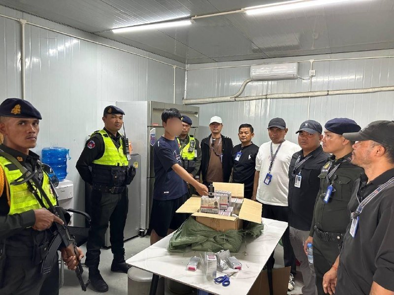 Armed personnel in uniforms and vests stand around a table displaying boxes of packaged items inside a prefabricated room.