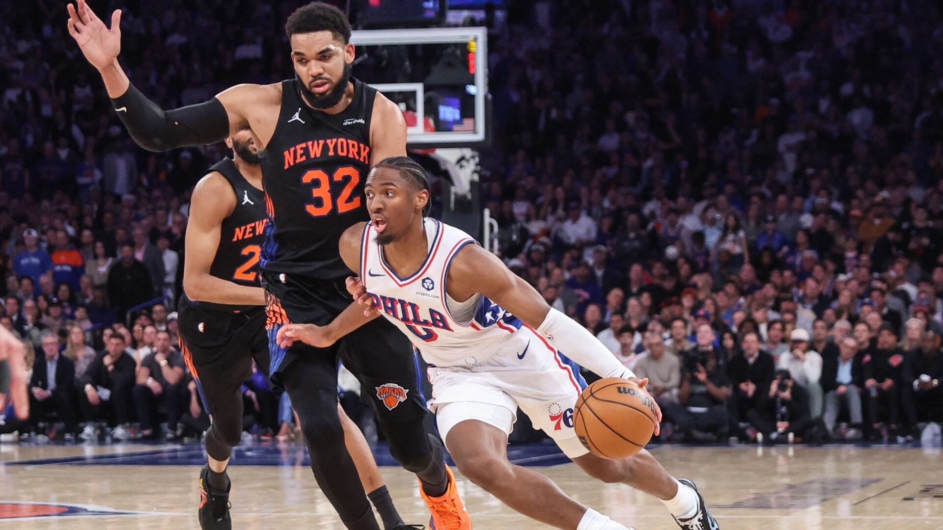 A Philadelphia 76ers player in a white jersey dribbles past a New York Knicks defender wearing number 32 during an NBA game before a crowded arena.