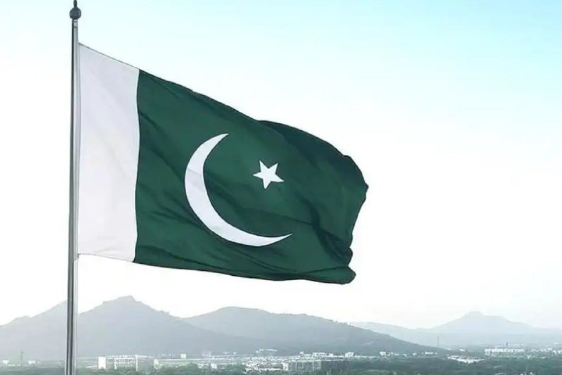 The green and white flag of Pakistan, featuring a crescent and star, waves on a flagpole against a hazy sky with distant mountains and a cityscape below.