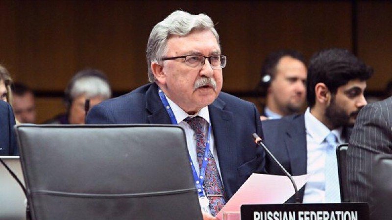 An older man with gray hair, glasses, and a mustache sits at a conference table behind a "RUSSIAN FEDERATION" placard, wearing a lanyard.