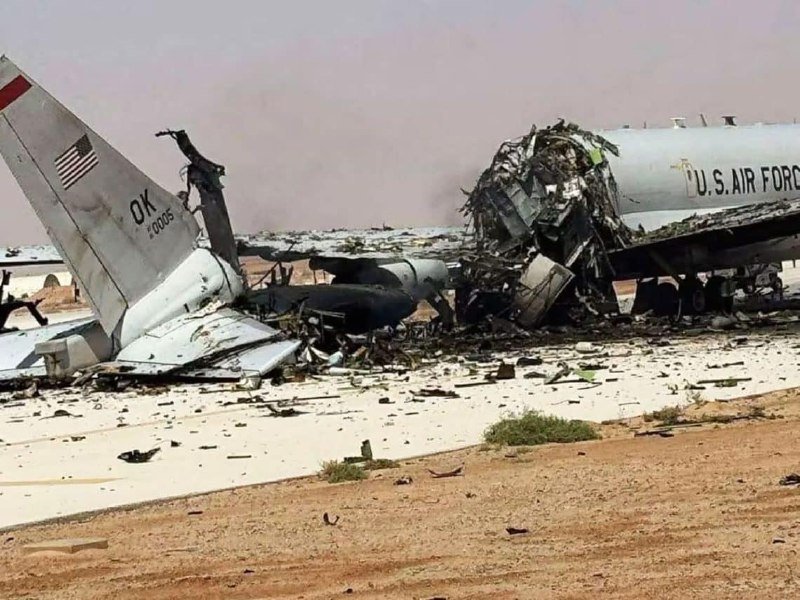 A destroyed U.S. Air Force aircraft marked "OK 2005" lies in wreckage across a paved runway, with debris scattered around its separated tail section under a hazy sky.