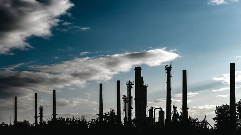 Silhouetted industrial smokestacks and refinery towers rise against a partly cloudy sky, with trees visible in the foreground.