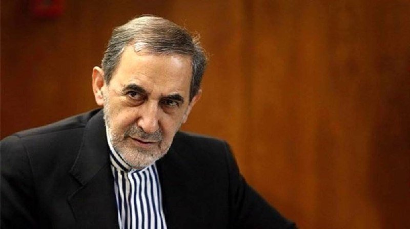 A gray-bearded man in a black suit jacket and striped shirt looks directly at the camera against a wooden panel background.