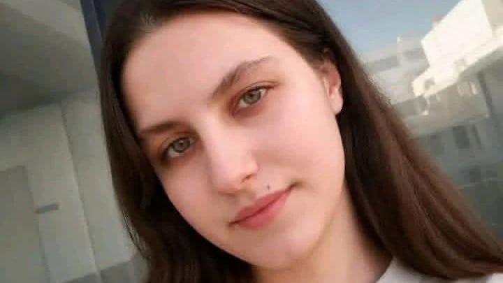 A close-up selfie of a woman with long brown hair, looking directly at the camera with a neutral expression, with a building visible in the background.