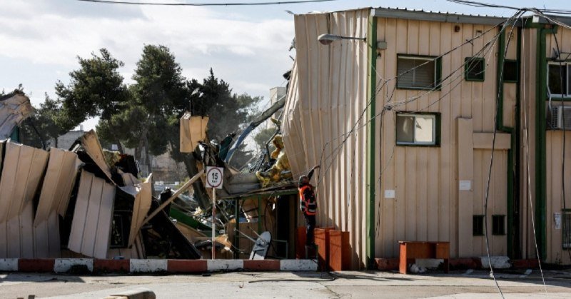 A worker in a high-visibility vest stands beside a partially collapsed metal building, with debris and twisted panels scattered in front of the damaged structure.