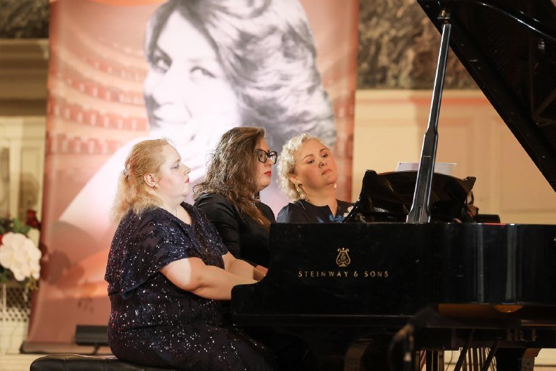 Three women perform at a Steinway & Sons grand piano on stage, with a large banner featuring a black-and-white portrait of a woman displayed behind them.