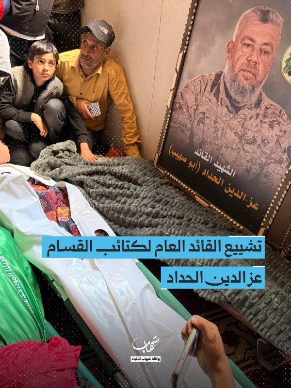 Mourners sit beside a shrouded body draped in a flag, next to a framed portrait of a bearded man with Arabic text, as overlaid Arabic captions identify the deceased.