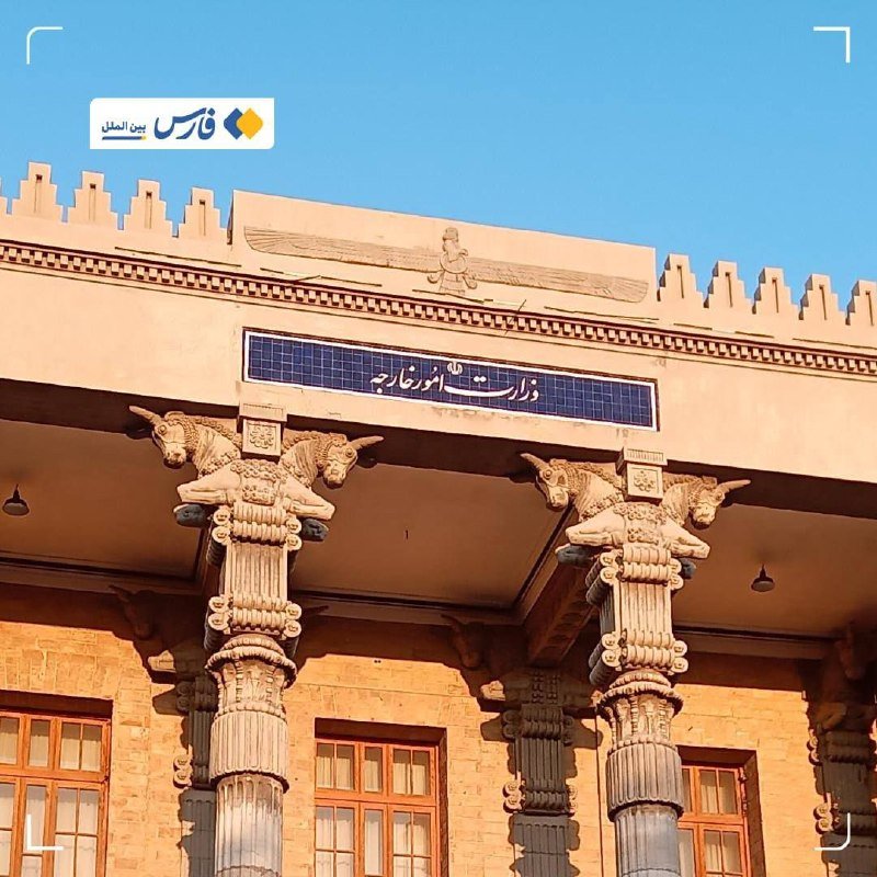 A sandstone building features ornate columns with carved bull capitals, a winged figure frieze, and a Persian sign reading "وزارت امور خارجه" against a clear blue sky.