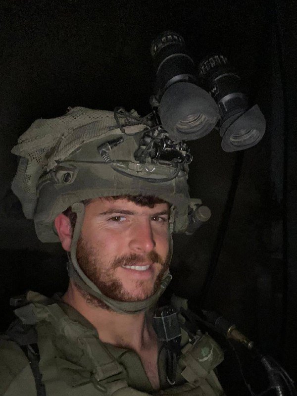 A bearded soldier in a tactical helmet with mounted night-vision goggles smiles at the camera in a dimly lit setting, wearing military gear.