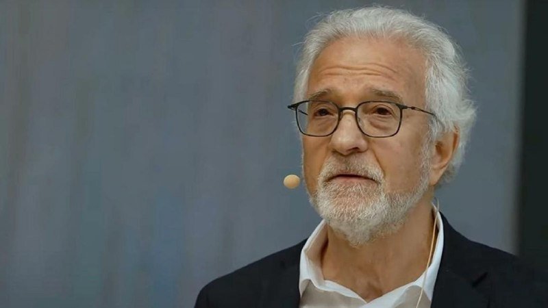 An older man with gray hair and beard, wearing glasses and a headset microphone, speaks against a gray background.