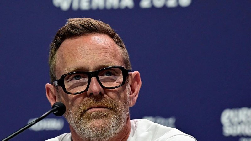 A bearded man with glasses speaks into a microphone against a blue backdrop displaying "Vienna 2020."