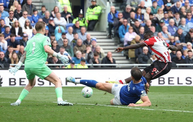 During a soccer match, a goalkeeper in green watches as a defender in blue slides on the grass to block a shot from an opposing player in a red and white striped jersey.