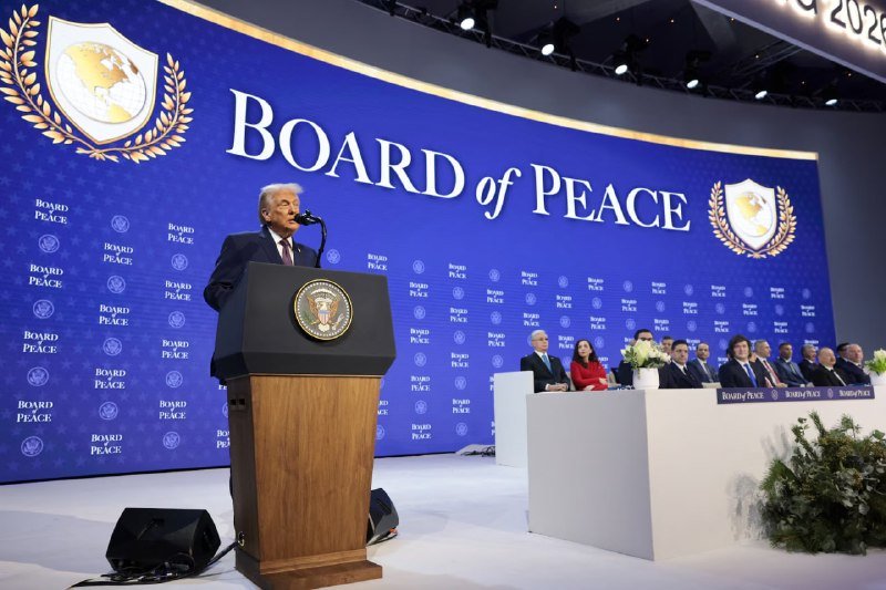 A man in a dark suit speaks at a podium bearing the presidential seal, backed by a "Board of Peace" display, while seated attendees observe from a nearby table.