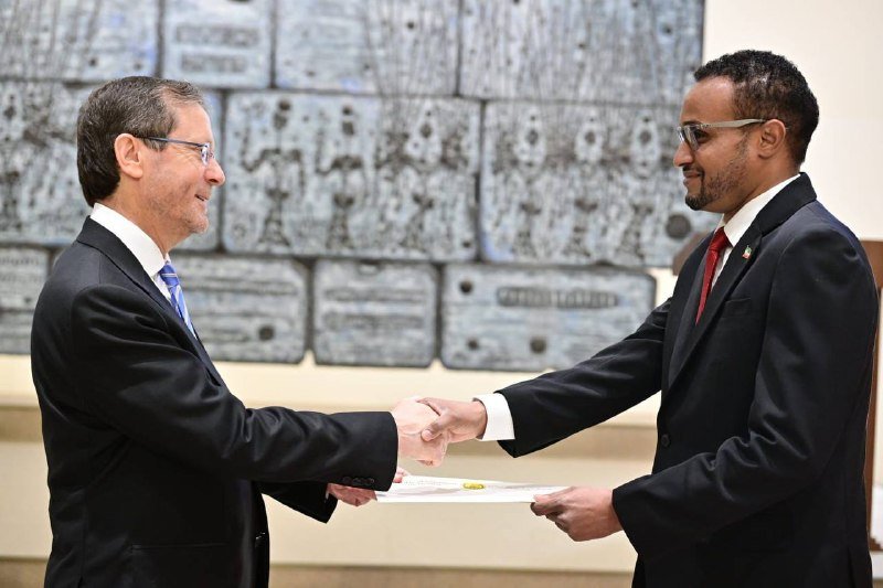Two men in suits shake hands and exchange a document during a formal indoor ceremony, standing before a decorative stone-like wall.