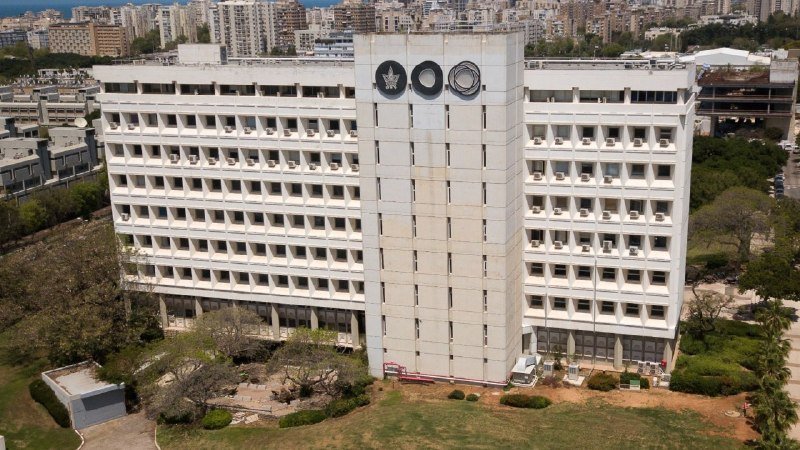 Aerial view of a large white multi-story building displaying three circular emblems on its facade, surrounded by greenery with a city skyline and sea in the background.
