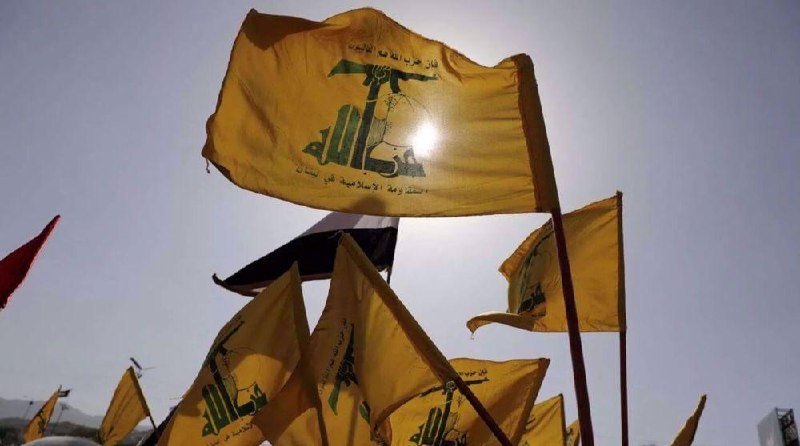 Multiple yellow flags bearing a green emblem and Arabic script wave against a bright, hazy sky, with one red flag partially visible on the left.