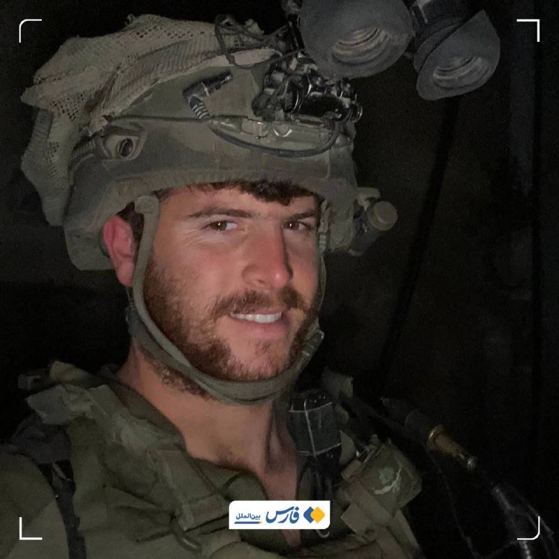 A bearded soldier wearing a combat helmet with mounted night-vision goggles smiles inside a dark vehicle, with a radio and tactical vest visible.