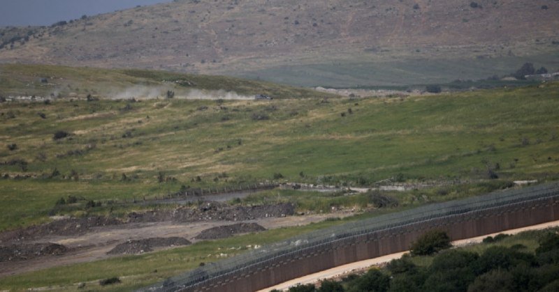 Dust rises near vehicles on a road behind a tall border fence in a grassy, hilly landscape.
