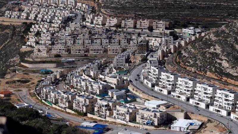Aerial view of densely packed white multi-story buildings climbing a rocky hillside, with winding roads and sparse vegetation visible.
