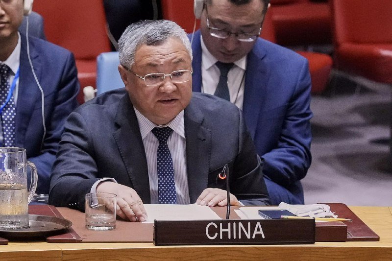 A gray-haired man in a dark suit and glasses speaks into a microphone at a desk marked "CHINA," with another suited man seated behind him wearing headphones.