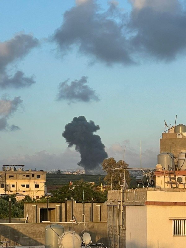 A large plume of dark smoke rises above a cityscape of low-rise buildings, water tanks, and satellite dishes under a partly cloudy sky.