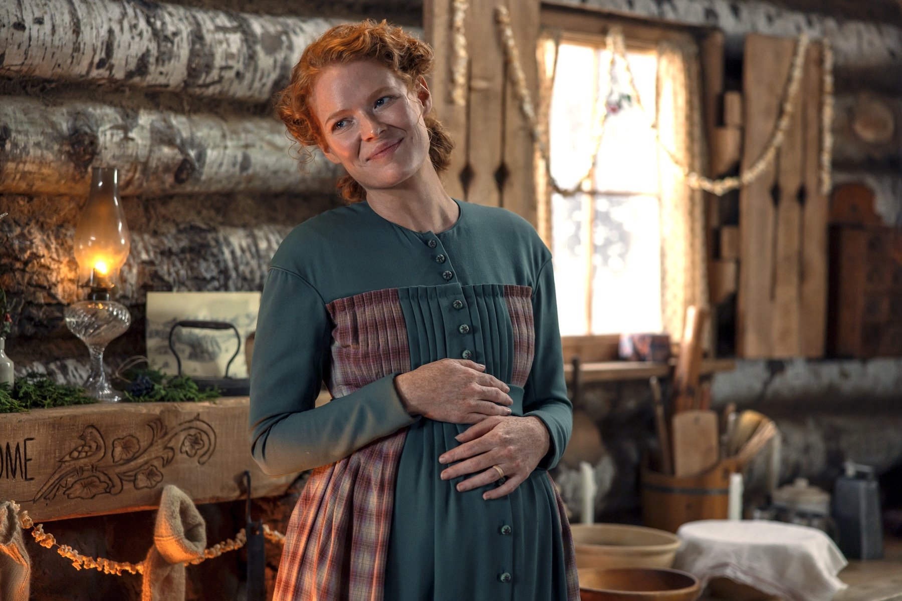 A red-haired woman in a green and plaid period dress stands smiling inside a rustic log cabin, her hands gently resting on her stomach.